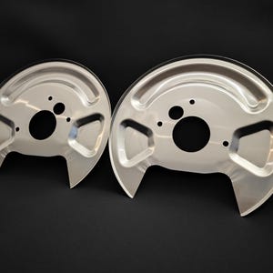 Puede incluir: Dos protectores contra el polvo de freno automotrices semicirculares plateados. Cada protector tiene un gran orificio central, orificios de montaje más pequeños y un borde curvo y elevado. Los protectores son de metal brillante sobre fondo negro.