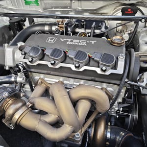 Op de afbeelding: Close-up van een automotor met een zwarte VTEC-T Honda-afdekking. De motor heeft meerdere spoelen, zilveren leidingen en een turbo. De motorruimte is zilver.