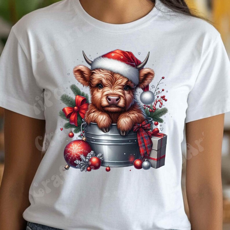 May include: White t-shirt featuring a festive illustration of a Highland cow calf wearing a Santa hat. The calf is in a metal bucket surrounded by Christmas ornaments, a red bow, and a wrapped gift. The design has a watercolor effect.