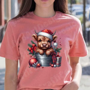 May include: A light red t-shirt featuring a festive graphic. The graphic depicts a brown Highland cow wearing a Santa hat, nestled in a metal bucket with Christmas decorations. Ornaments, a red bow, and a wrapped gift complete the holiday scene.