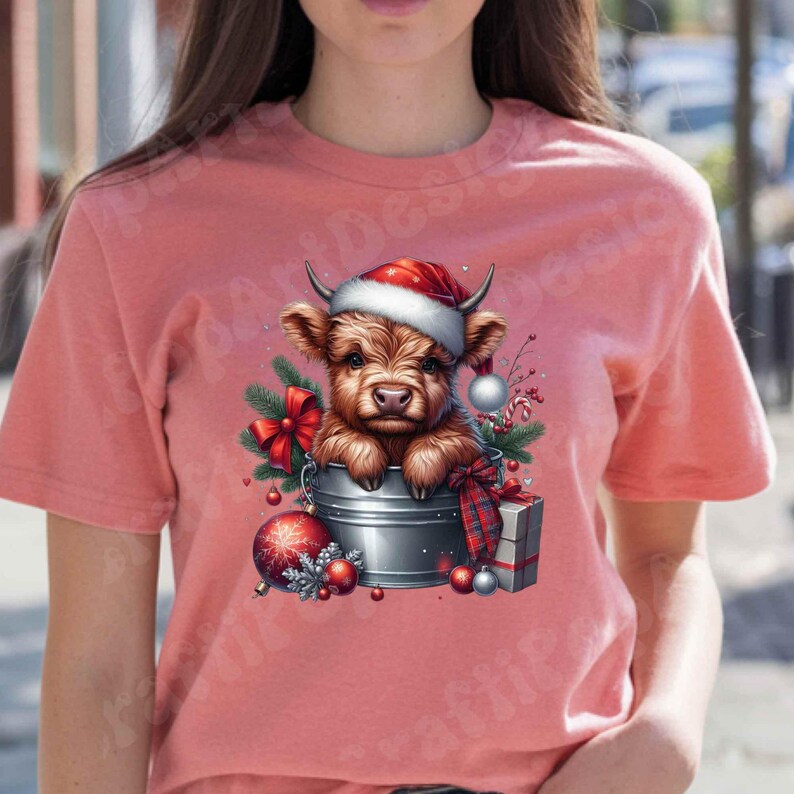 May include: A light red t-shirt featuring a festive graphic. The graphic depicts a brown Highland cow wearing a Santa hat, nestled in a metal bucket with Christmas decorations. Ornaments, a red bow, and a wrapped gift complete the holiday scene.