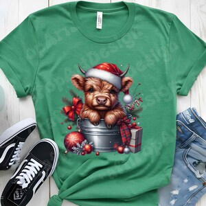 May include: Green t-shirt featuring a festive illustration of a Highland cow calf wearing a Santa hat, nestled in a metal bucket with Christmas decorations. The design includes a red bow, ornaments, and a wrapped gift. Black and white sneakers are visible.