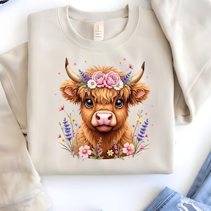 May include: A beige sweatshirt with a brown and white Highland cow wearing a flower crown. The flowers are pink, white, and purple.