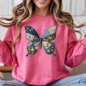 May include: A pink sweatshirt featuring a large butterfly design. The butterfly has patchwork wings in shades of blue, green, and beige, adorned with floral accents. The sweatshirt is a solid pink color.