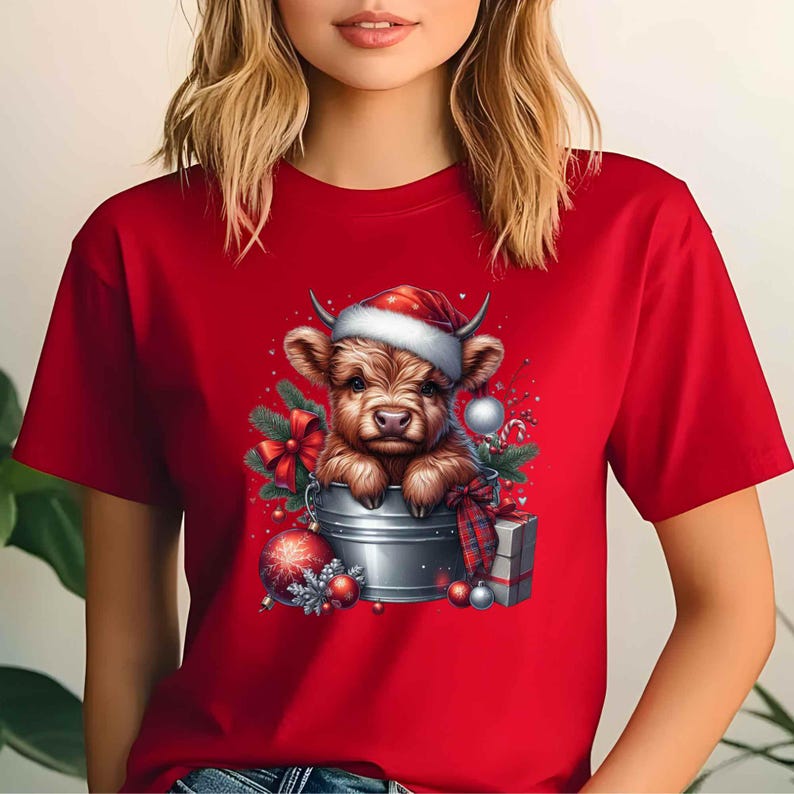 May include: Red t-shirt featuring a festive illustration of a Highland cow wearing a Santa hat, nestled in a metal bucket. The design includes Christmas ornaments, a plaid bow, and a wrapped gift, creating a holiday theme.