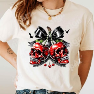 May include: Cream-colored t-shirt featuring a graphic of two red skull cherries with a black bow and bats. The cherries have a lace trim and small red cherries. A unique design for a spooky aesthetic.