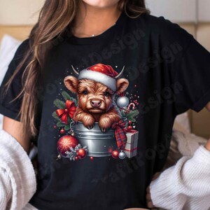 May include: Black t-shirt featuring a festive graphic of a Highland cow wearing a Santa hat, emerging from a metal bucket. The design includes Christmas ornaments, a red bow, and a wrapped gift, creating a holiday theme.
