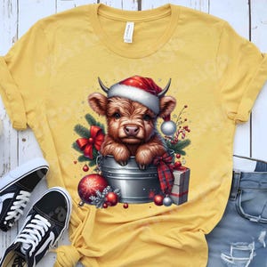 May include: Yellow t-shirt featuring a festive graphic of a Highland cow wearing a Santa hat, nestled in a metal bucket. The design includes Christmas ornaments, a red bow, and a wrapped gift, creating a holiday theme.