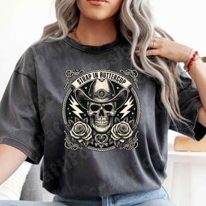 May include: Dark gray t-shirt featuring a circular graphic with a skull wearing a cowboy hat. The graphic includes roses, lightning bolts, and the text "STRAP IN BUTTERCUP" in a banner. The design is in black and white.