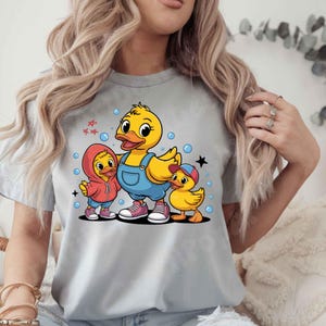 May include: A light gray t-shirt featuring a cartoon illustration of three yellow ducks. The central duck wears blue overalls, while the other two wear a red hoodie and a blue cap. Bubbles and stars surround the ducks.