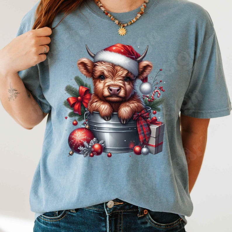 May include: A light blue t-shirt featuring a festive illustration. A brown Highland cow wearing a Santa hat is nestled in a metal bucket, surrounded by Christmas ornaments, a red bow, and a wrapped gift. The design evokes a holiday theme.