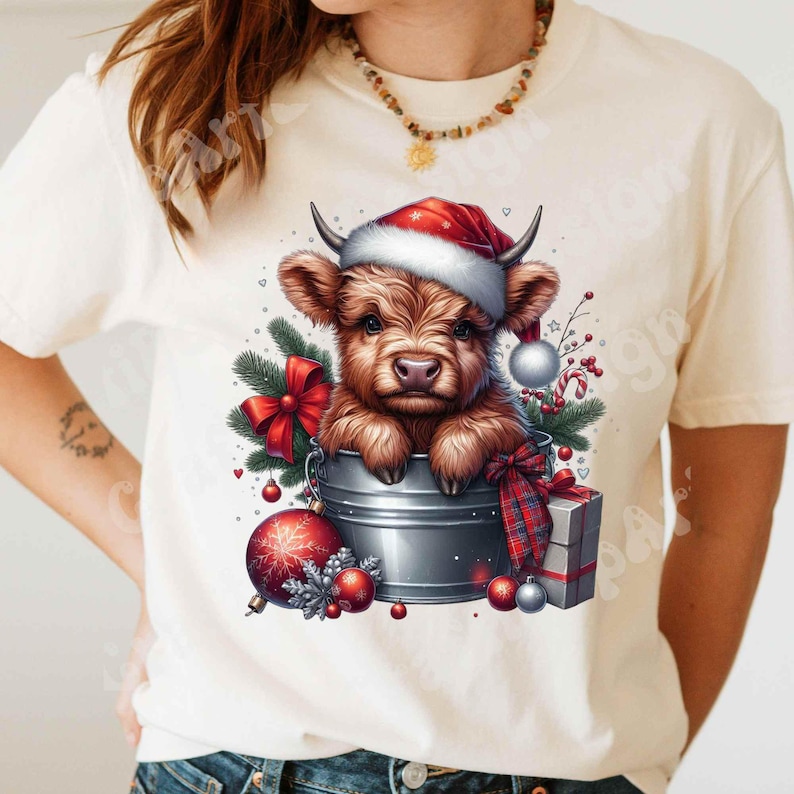 May include: Cream-colored t-shirt featuring a festive illustration. A brown Highland cow wearing a Santa hat peeks out of a metal bucket, surrounded by Christmas ornaments, a red bow, and a wrapped gift. The design has a watercolor effect.