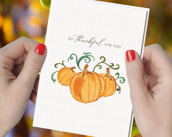 May include: A white greeting card featuring a watercolor illustration of three orange pumpkins with green vines. The text "SO THANKFUL FOR YOU" is printed in a dark brown script font above the pumpkins on the front of the folded card.