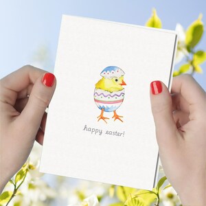 May include: A white greeting card featuring a watercolor illustration of a yellow chick hatching from a decorated egg with blue, pink, and green patterns. The text "happy easter!" is printed in a simple, dark gray font below the chick.