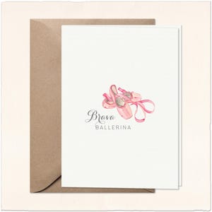 May include: A white greeting card featuring a watercolor illustration of pink ballet slippers with ribbons. The text Brava BALLERINA is printed in black below the slippers. The card is placed on top of a brown kraft paper envelope.