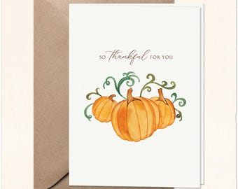 May include: A white greeting card featuring a watercolor illustration of three orange pumpkins with green vines and the text "SO THANKFUL FOR YOU" in a dark brown script font, accompanied by a plain brown envelope positioned behind the card.