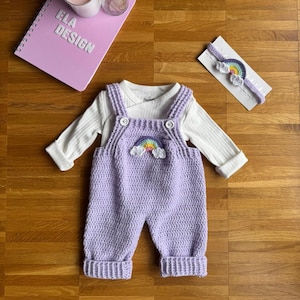 May include: A purple crocheted baby overall with a rainbow and cloud applique. The overall has a white long-sleeved shirt underneath. A matching rainbow headband is also included.