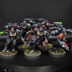 Puede incluir: Un grupo de cinco figuras en miniatura de marines espaciales pintadas con armadura negra con detalles en rojo, emblemas dorados en el pecho y mochilas negras. Cada figura está montada sobre una base circular con suelo texturizado.