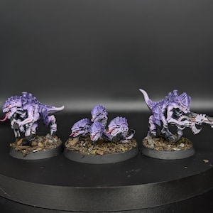 May include: Three miniature alien creature figurines with purple and white skin, sharp teeth, and pink tongues. Two larger figures stand on the sides, one holding a weapon, while a smaller cluster of three heads sits on a base in the center.