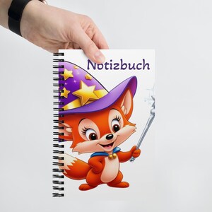 Op de afbeelding: Een spiraalgebonden notitieboek met een kleurrijke illustratie van een vos die een paarse en gele tovenaarshoed draagt en een toverstaf vasthoudt. De tekst "Notizbuch" staat op de omslag.