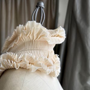 Puede incluir: Un cuello de tela plisada de color crema con un borde con volantes, exhibido en un maniquí. El cuello tiene un diseño estructurado y en capas, y es probablemente un accesorio de moda. El fondo presenta cortinas de tonos neutros.