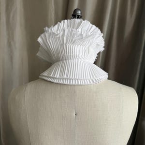 May include: A white, pleated collar, possibly for historical or theatrical use, is shown on a mannequin. The collar has multiple layers of finely pleated fabric, creating a ruffled effect around the neck. The collar is a bright white colour.