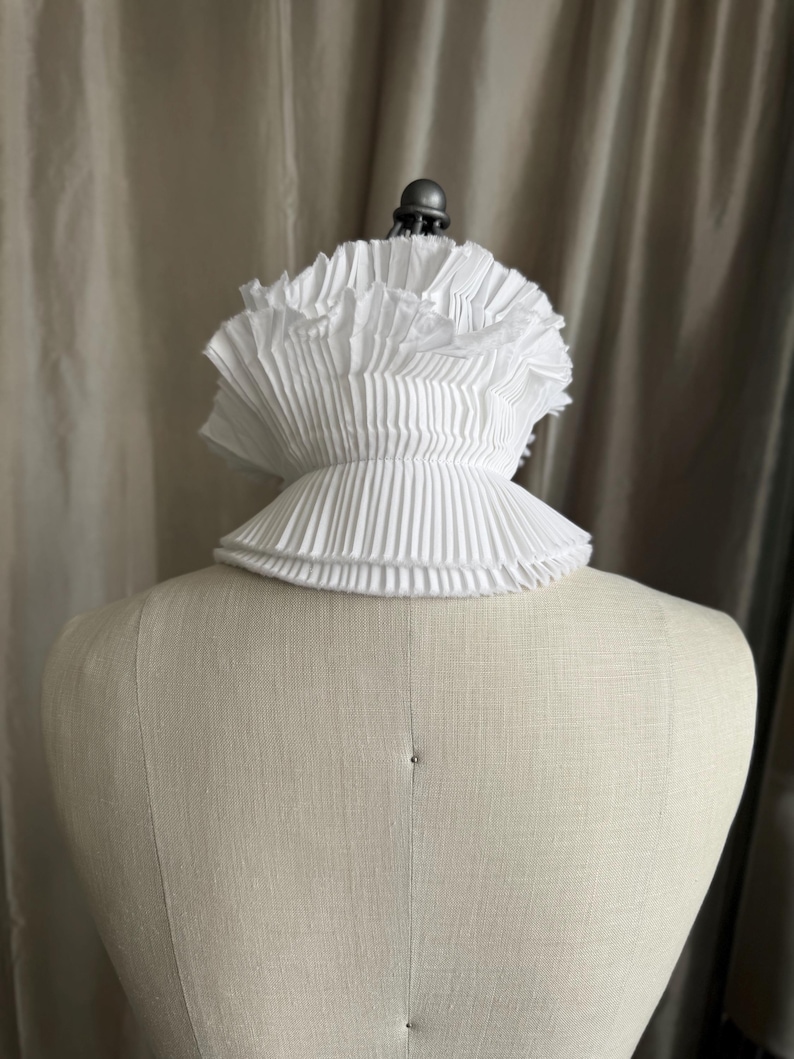 May include: A white, pleated collar, possibly for historical or theatrical use, is shown on a mannequin. The collar has multiple layers of finely pleated fabric, creating a ruffled effect around the neck. The collar is a bright white colour.