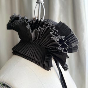 May include: A black, pleated collar with a ruffled edge and a ribbon tie. The collar is made of a dark fabric and is displayed on a mannequin. The design is reminiscent of historical fashion.