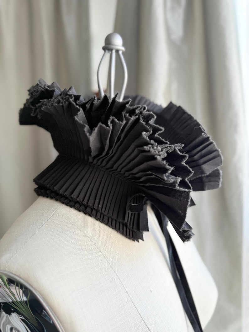 May include: A black, pleated collar with a ruffled edge and a ribbon tie. The collar is made of a dark fabric and is displayed on a mannequin. The design is reminiscent of historical fashion.