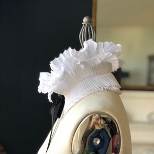 Puede incluir: Un cuello blanco con volantes y una pajarita negra. El cuello está en un maniquí blanco con una superficie reflectante.