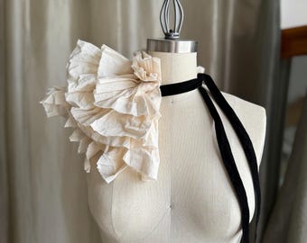 May include: A cream-colored, ruffled shoulder piece with a black velvet ribbon, displayed on a mannequin. The ruffles are layered and textured, creating a voluminous effect. The black ribbon is tied around the neck and hangs down.