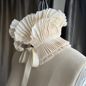 May include: A cream-coloured, pleated collar with a ruffled edge and a satin ribbon tie. The collar is displayed on a mannequin, highlighting its delicate texture and design. A vintage-inspired accessory.