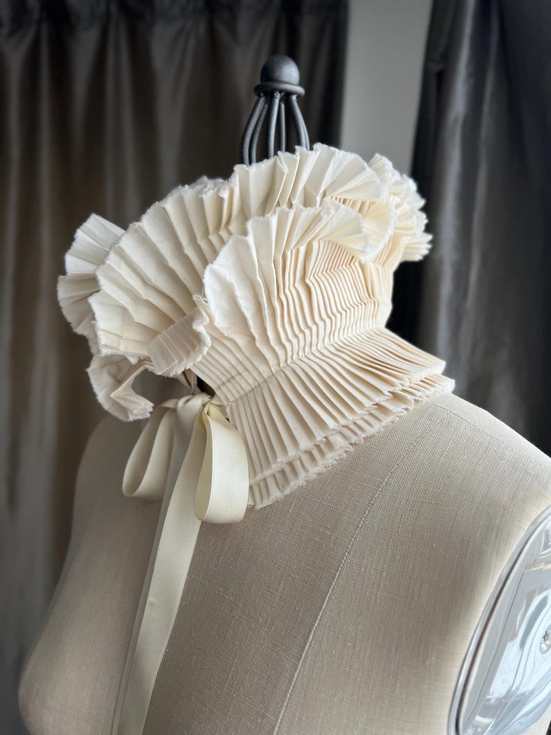 May include: A cream-coloured, pleated collar with a ruffled edge and a satin ribbon tie. The collar is displayed on a mannequin, highlighting its delicate texture and design. A vintage-inspired accessory.