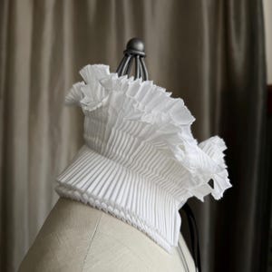 May include: A white, pleated collar with a ruffled edge, displayed on a mannequin. The collar has a structured, layered design and is made of a crisp fabric. A black ribbon hangs from the back.