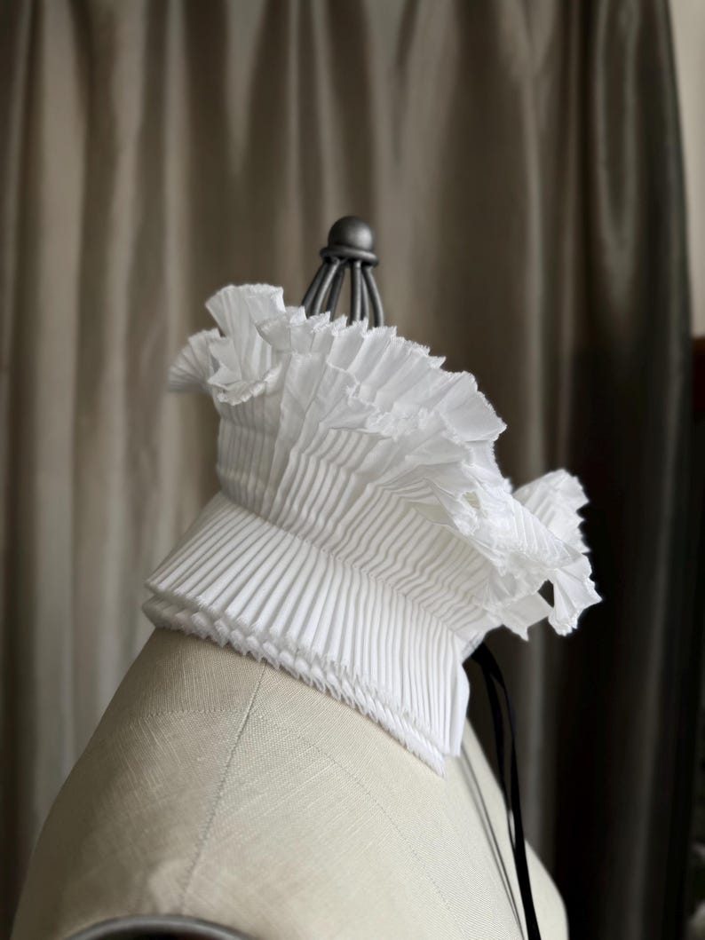 May include: A white, pleated collar with a ruffled edge, displayed on a mannequin. The collar has a structured, layered design and is made of a crisp fabric. A black ribbon hangs from the back.