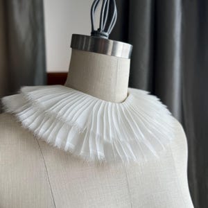 May include: A white, ruffled collar made of fabric. The collar is displayed on a white dress form.