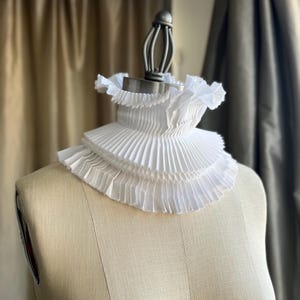May include: A white, pleated Elizabethan ruff collar, displayed on a dress form. The collar features multiple layers of finely pleated fabric, creating a ruffled effect around the neck. The dress form is a neutral beige colour, set against a blurred background.