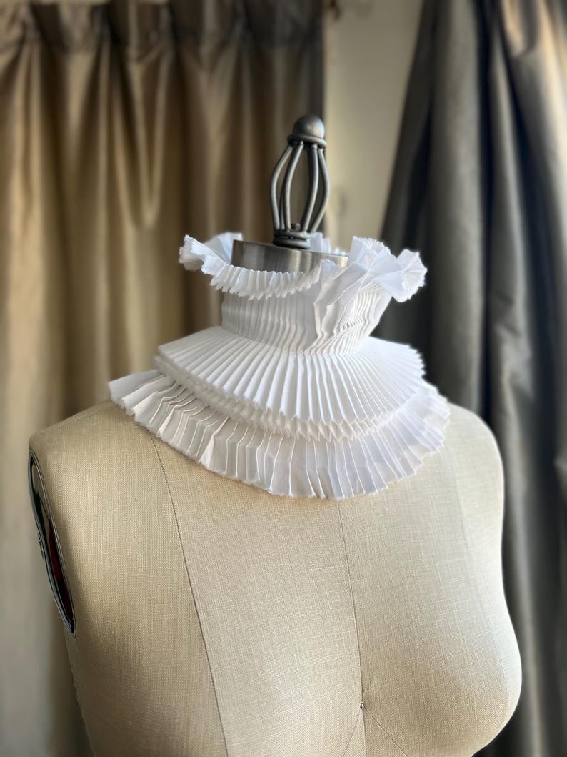 May include: A white, pleated Elizabethan ruff collar, displayed on a dress form. The collar features multiple layers of finely pleated fabric, creating a ruffled effect around the neck. The dress form is a neutral beige colour, set against a blurred background.