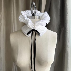 May include: A white, pleated ruffle collar with a black satin ribbon tie, displayed on a mannequin. The collar has a layered, textured appearance, and the ribbon hangs down the front. The background features neutral-toned curtains.