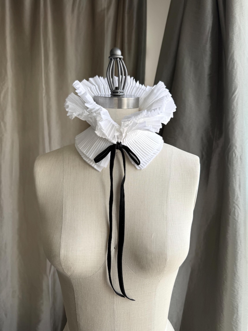 May include: A white, pleated ruffle collar with a black satin ribbon tie, displayed on a mannequin. The collar has a layered, textured appearance, and the ribbon hangs down the front. The background features neutral-toned curtains.