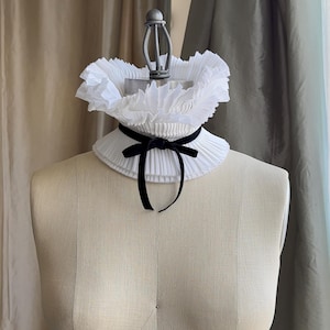 May include: A white, pleated collar with a black velvet ribbon bow, displayed on a mannequin. The collar has a ruffled edge and a classic design. The mannequin is a neutral beige colour, and the background features neutral-toned curtains.