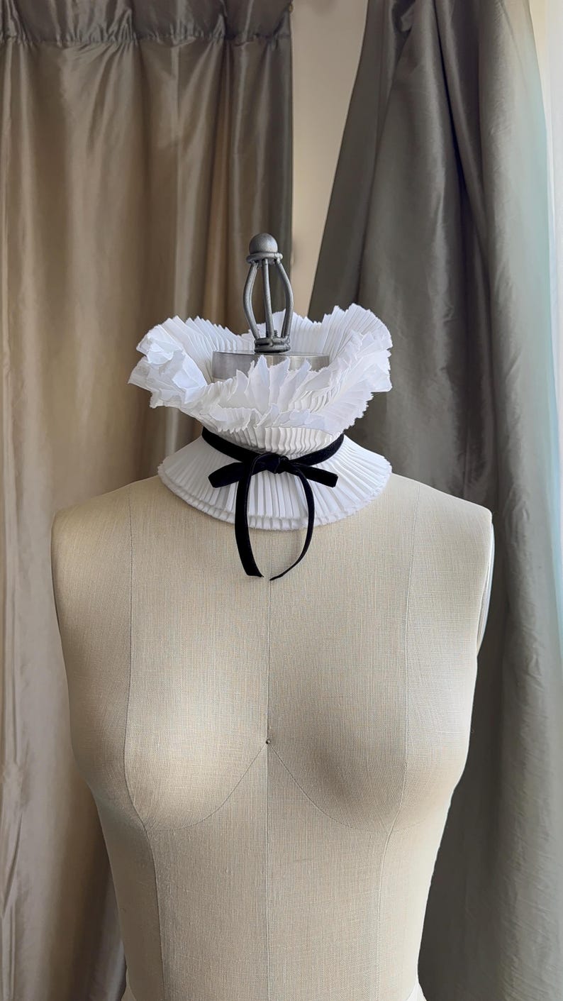 May include: A white, pleated collar with a black velvet ribbon bow, displayed on a mannequin. The collar has a ruffled edge and a classic design. The mannequin is a neutral beige colour, and the background features neutral-toned curtains.