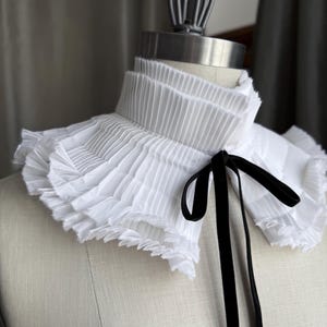 May include: A white pleated collar with a ruffled edge and a black velvet ribbon tie. The collar is displayed on a mannequin, showcasing its layered design and texture. The style is reminiscent of vintage fashion.