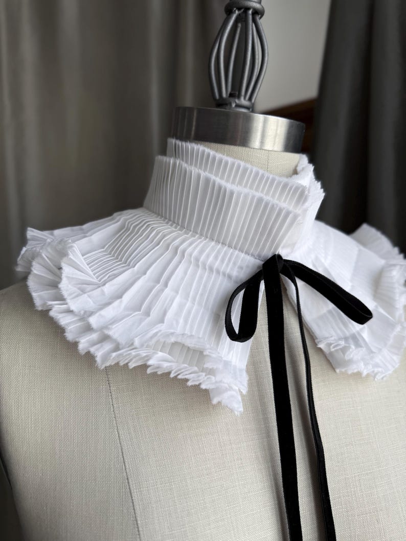 May include: A white pleated collar with a ruffled edge and a black velvet ribbon tie. The collar is displayed on a mannequin, showcasing its layered design and texture. The style is reminiscent of vintage fashion.