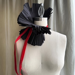 May include: A black pleated fabric neck ruff accessory featuring a structured, fan-like design that encircles the neck. The piece is secured with a thin, bright red velvet ribbon that hangs down in two long, loose tails against the torso.