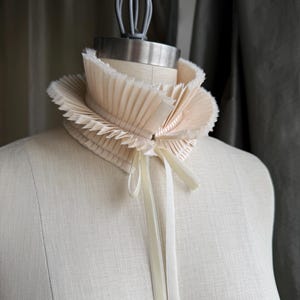 Könnte beinhalten: Eine cremefarbene, plissierte Halskrause aus Stoff mit einem hohen, stehenden Kragen und ausgefransten Kanten. Das Accessoire wird vorne mit zwei langen, passenden cremefarbenen Bändern befestigt, die vertikal von der Mitte herabhängen.