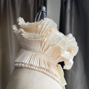 May include: A cream-coloured, pleated fabric collar on a mannequin. The collar has multiple layers of finely pleated material, creating a ruffled effect. The design is elegant and detailed, with a soft, flowing appearance. The collar is a fashion accessory.