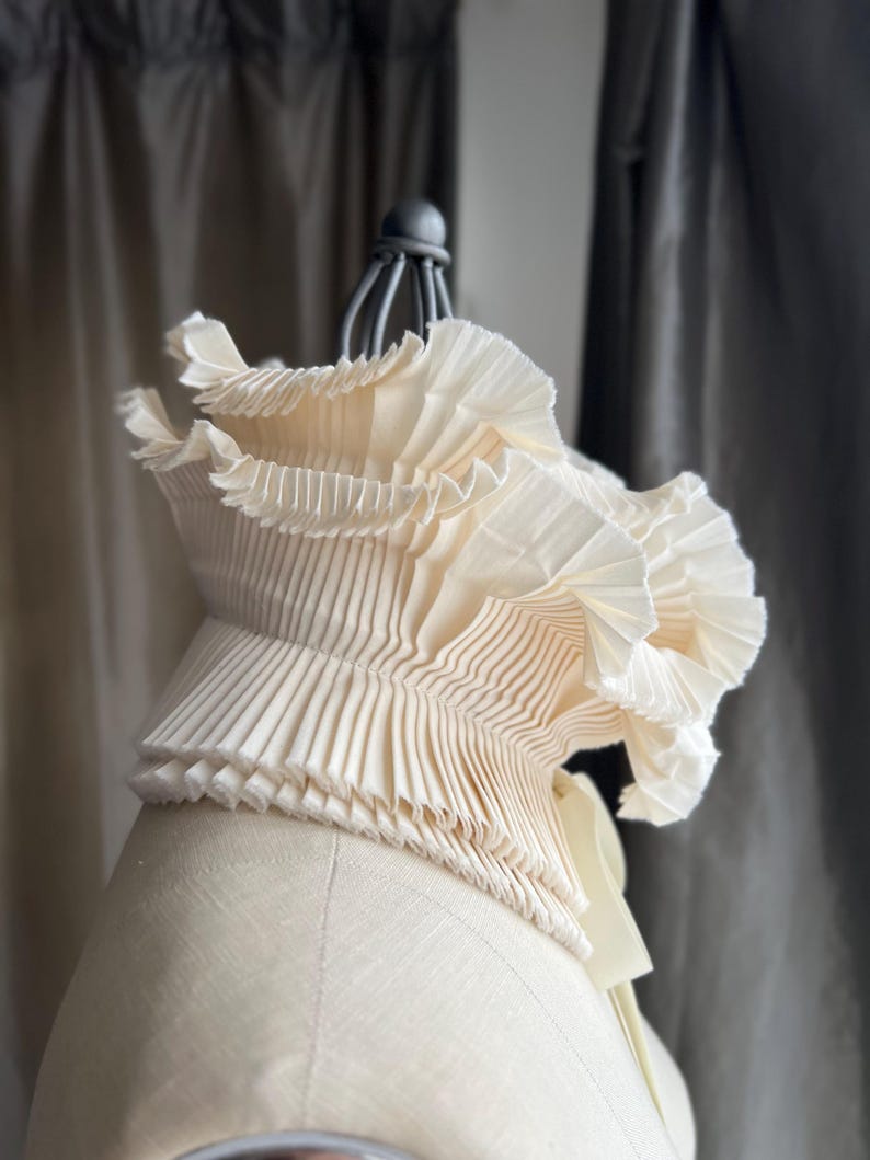 May include: A cream-coloured, pleated fabric collar on a mannequin. The collar has multiple layers of finely pleated material, creating a ruffled effect. The design is elegant and detailed, with a soft, flowing appearance. The collar is a fashion accessory.