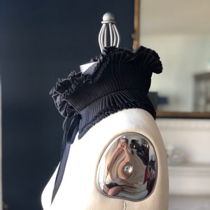 May include: A black pleated collar with a ruffled edge and a satin ribbon tie. The collar is displayed on a mannequin with a silver shoulder cap. The background is a dark blue wall and a mirror.