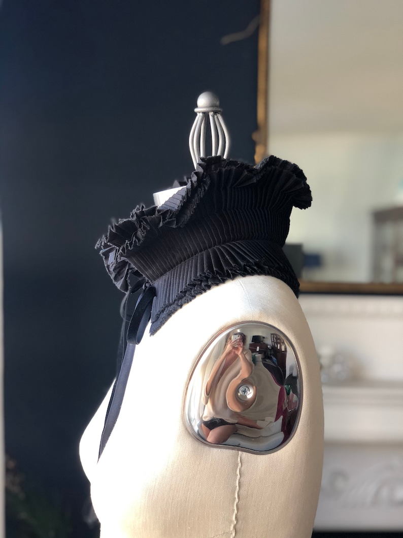 May include: A black pleated collar with a ruffled edge and a satin ribbon tie. The collar is displayed on a mannequin with a silver shoulder cap. The background is a dark blue wall and a mirror.